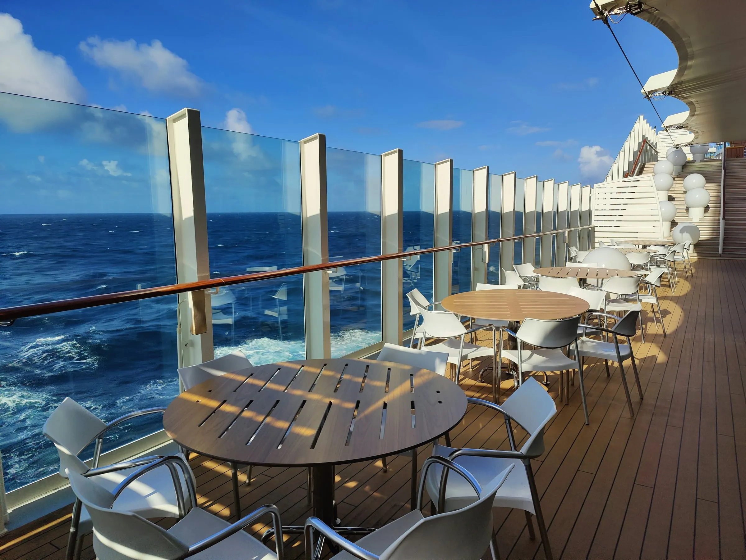 Leere Tische und Stühle auf einem Schiffsdeck hinter einer Glasreling, dahinter das offene Meer