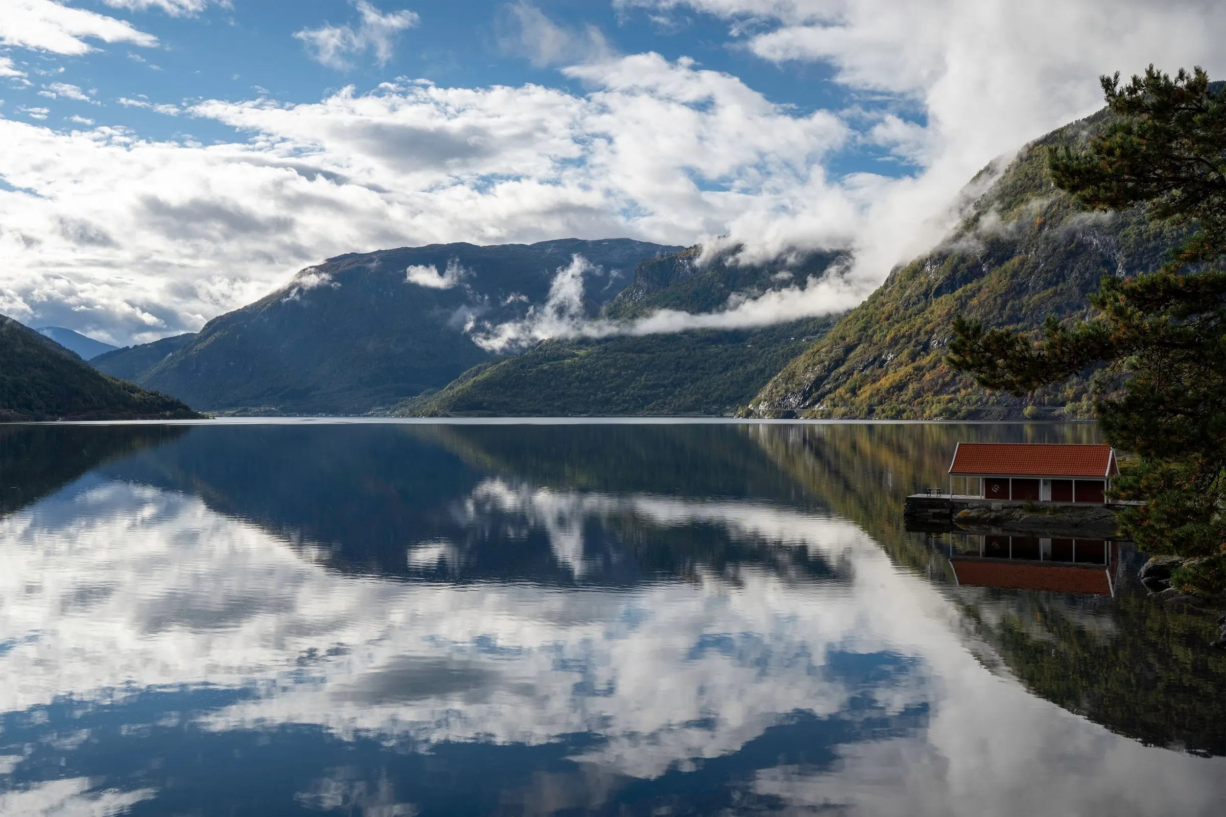 Ruhiger Fjord zwischen bewaldeten Bergen, Wolken spiegeln sich im Wasser, am Ufer steht ein rotes Bootshaus